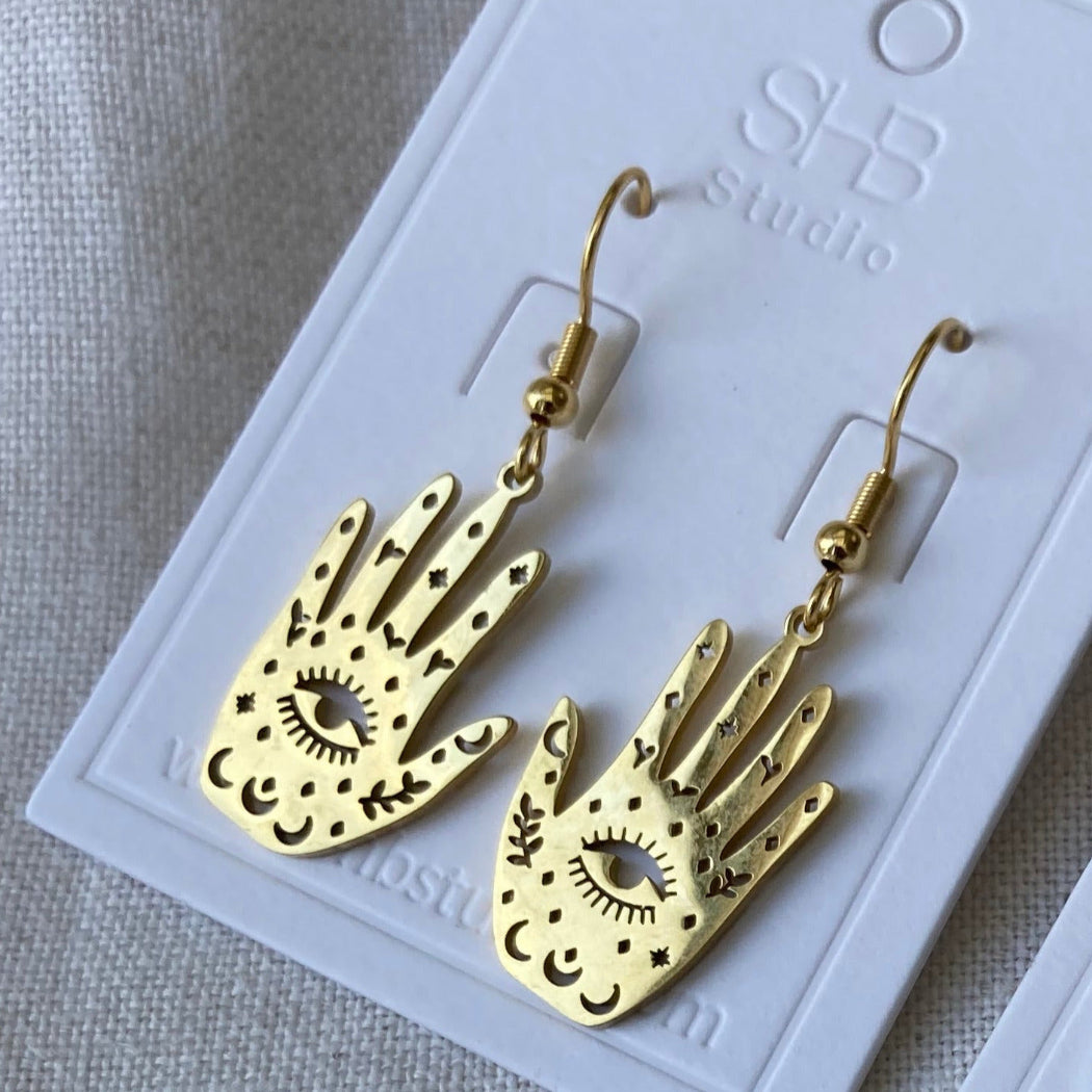 A pair of gold-plated stainless steel earrings with a hand and third eye design, displayed on a white card with 'SHB studio' branding.
