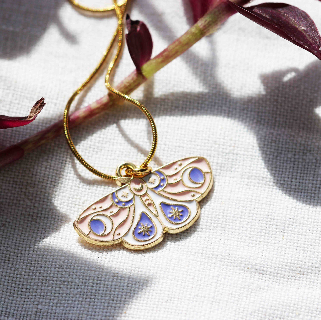 A gold-colored butterfly necklace with a stainless steel snake chain and a zinc alloy butterfly pendant, displayed on a white fabric surface with a shadow of a plant in the background.