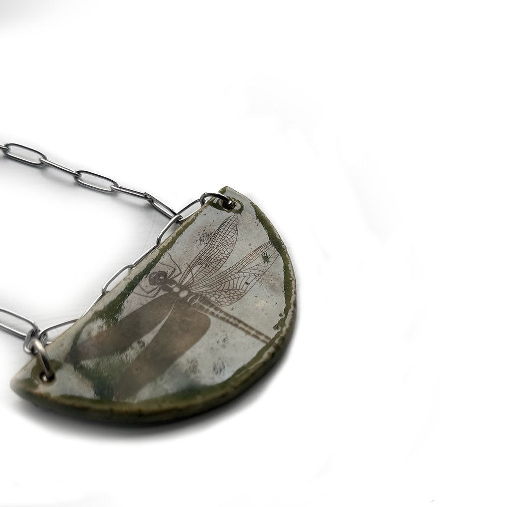 Handmade half-moon ceramic pendant necklace with a dragonfly on a stainless steel paperclip chain white background
