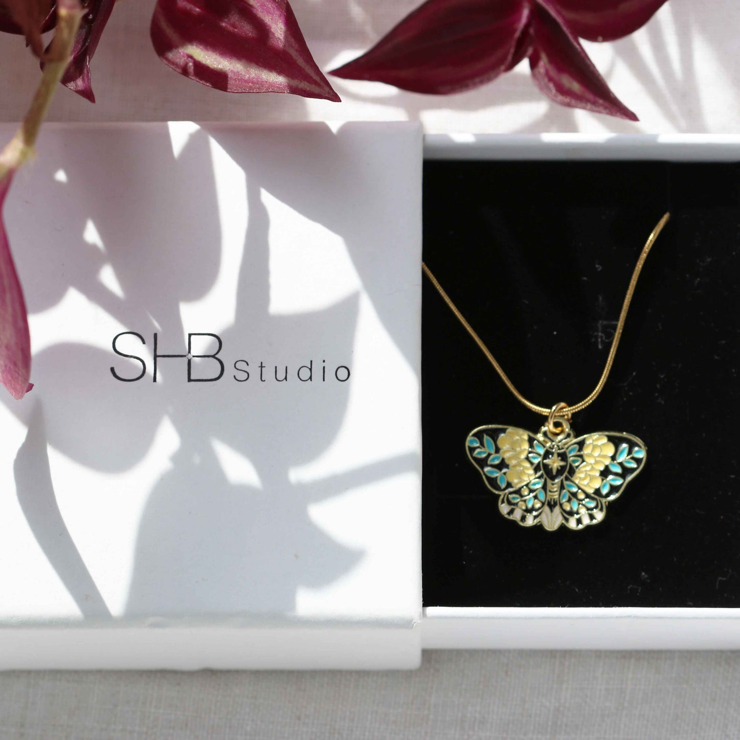 Butterfly necklace in a box with SHB Studio branding, surrounded by pink flowers.