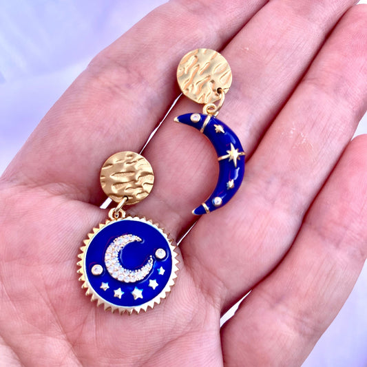 Blue and gold earrings with celestial design held in a hand against a light purple background