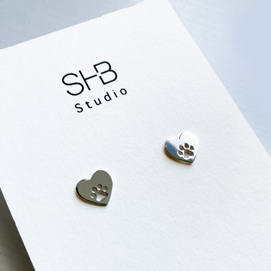 A pair of sterling silver heart and paw stud earrings placed on a white card with 'SHB Studio' printed on it.