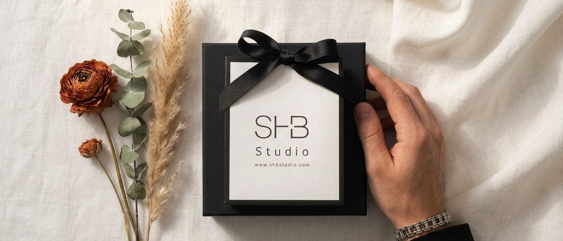 A black gift box with a black ribbon bow and a white insert card printed with 'SHB Studio' is held by a person's hand on a white fabric background, next to dried flowers and greenery.