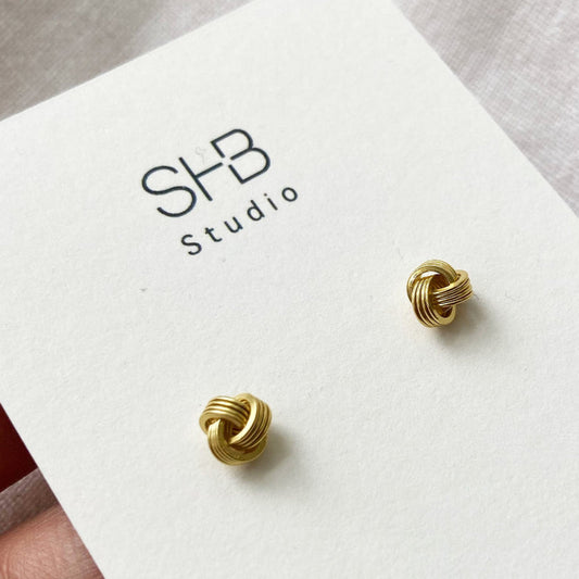 A pair of gold-plated knot stud earrings on a white card with 'SHB Studio' branding.