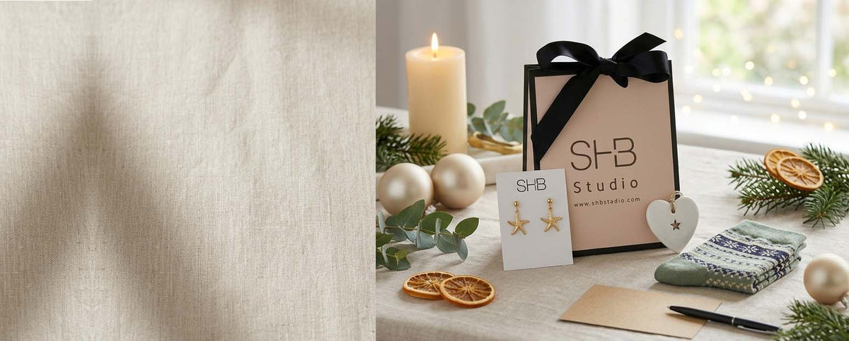A styled holiday gift scene featuring an SHB Studio paper shopping bag with a black ribbon, a matching SHB Studio card, gold star-shaped earrings, dried orange slices, pine branches, a candle, and a knitted scarf on a table.