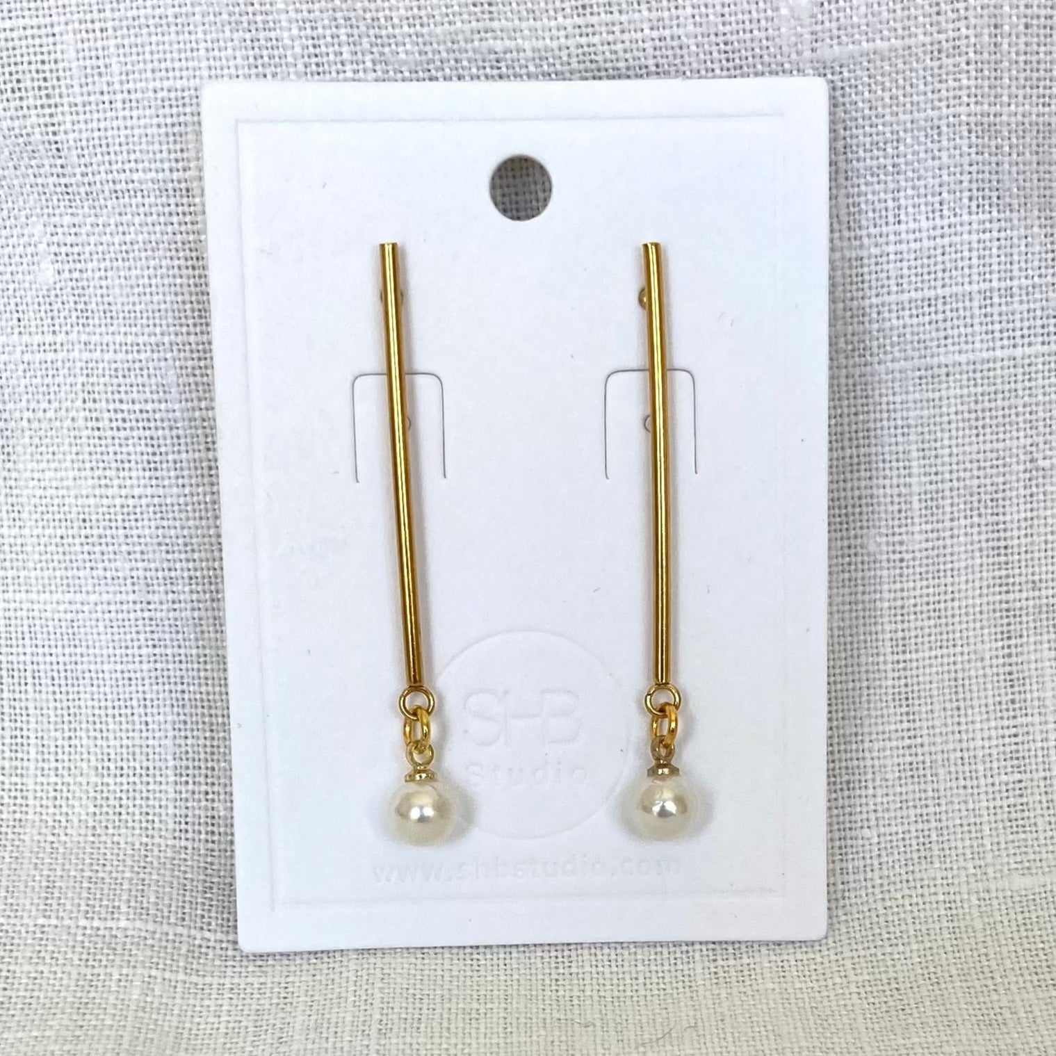A pair of minimal long gold and pearl earrings displayed on a white card, with the pearl drops at the bottom of gold-plated rods.