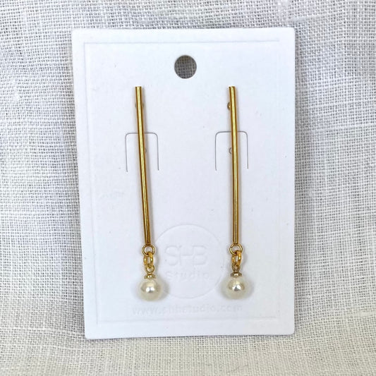 A pair of minimal long gold and pearl earrings displayed on a white card, with the pearl drops at the bottom of gold-plated rods.