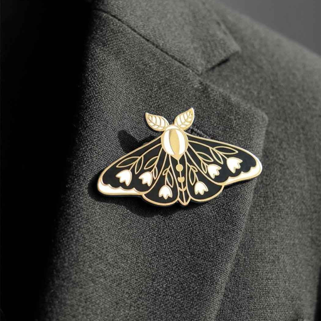 A gold and black moth-shaped lapel pin with white floral details, pinned to a dark charcoal gray suit jacket lapel.