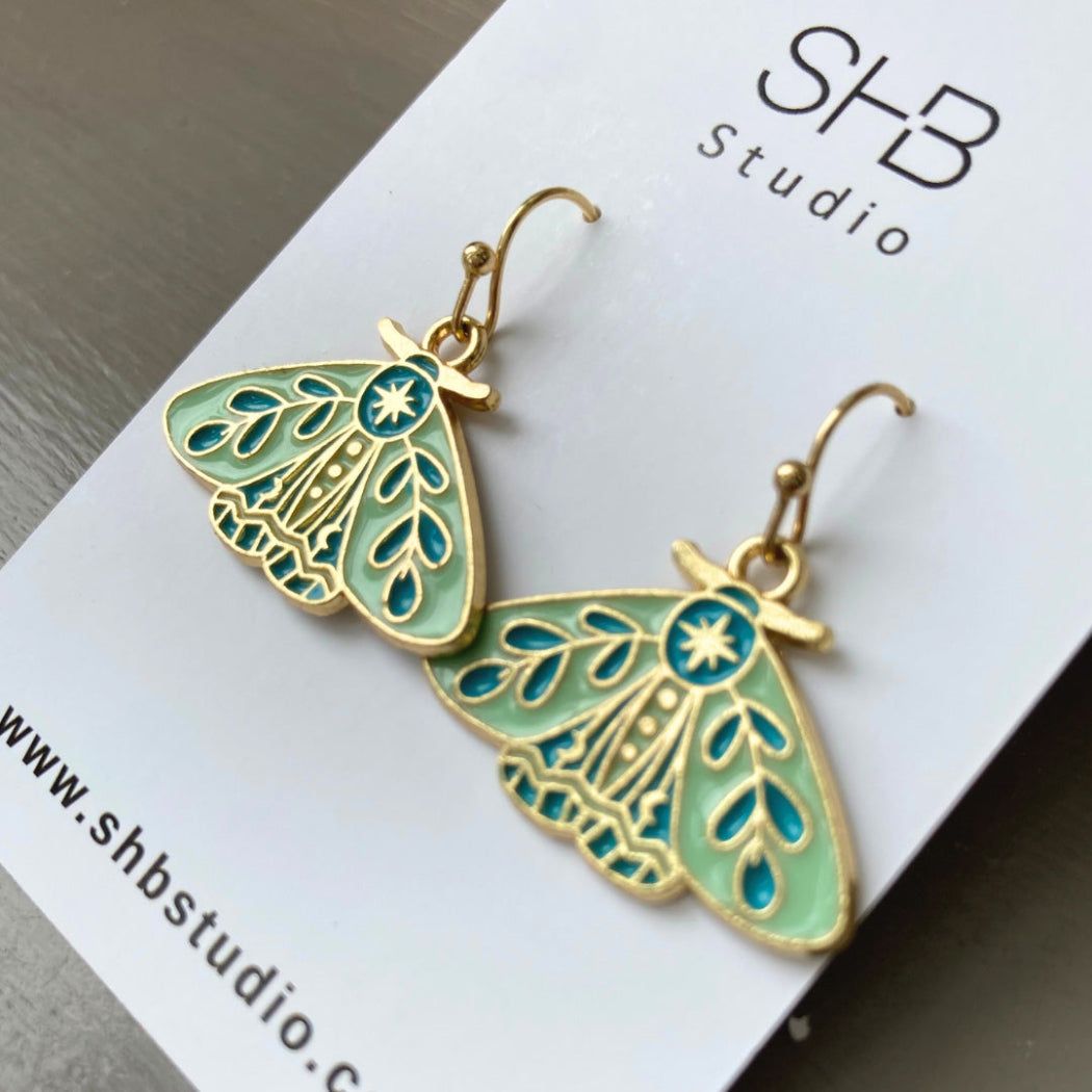 A pair of turquoise and gold moth design hook earrings on a white background, with 'SHB Studio' written above.