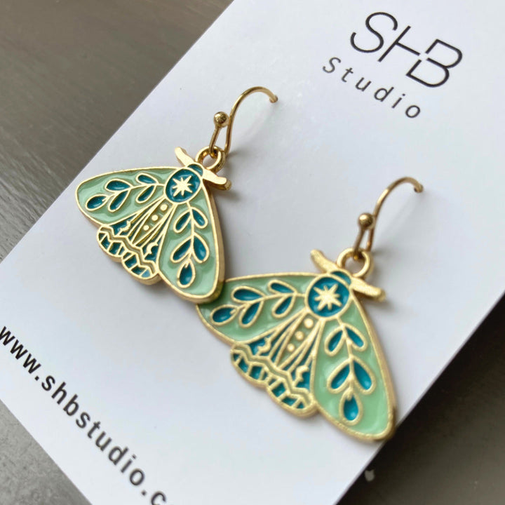 A pair of turquoise and gold moth design hook earrings on a white background, with &
