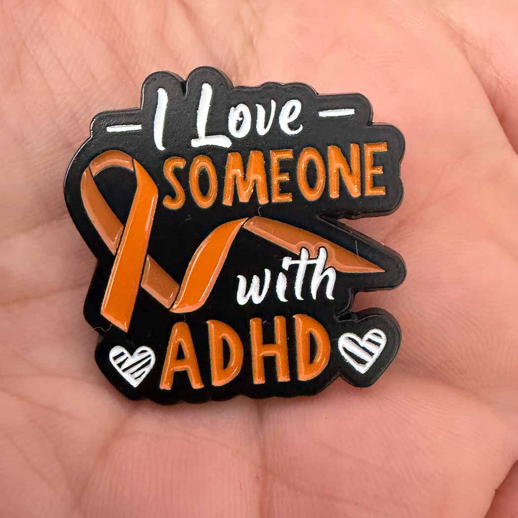A close-up of a hand holding a black enamel pin that reads &