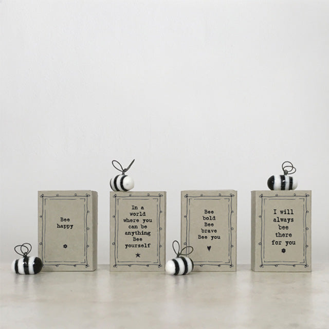 Four decorative rectangular boxes with printed bee-themed quotes are arranged on a light surface against a plain white background, with small striped hanging ornaments placed around them.