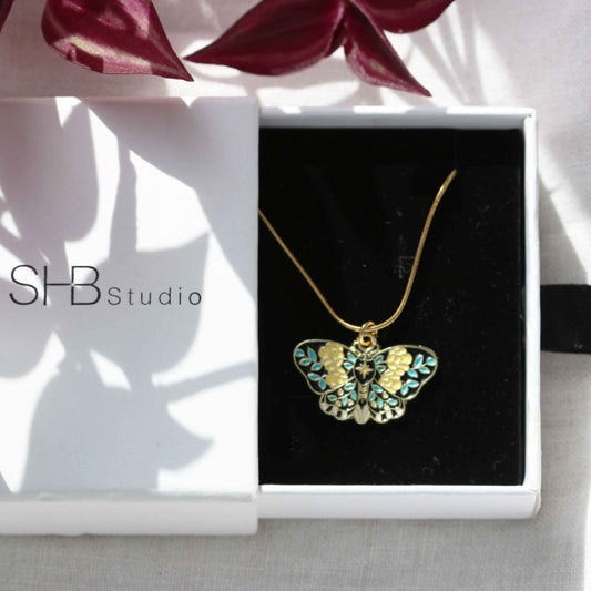 Butterfly necklace in a box with SHB Studio branding, surrounded by pink flowers.