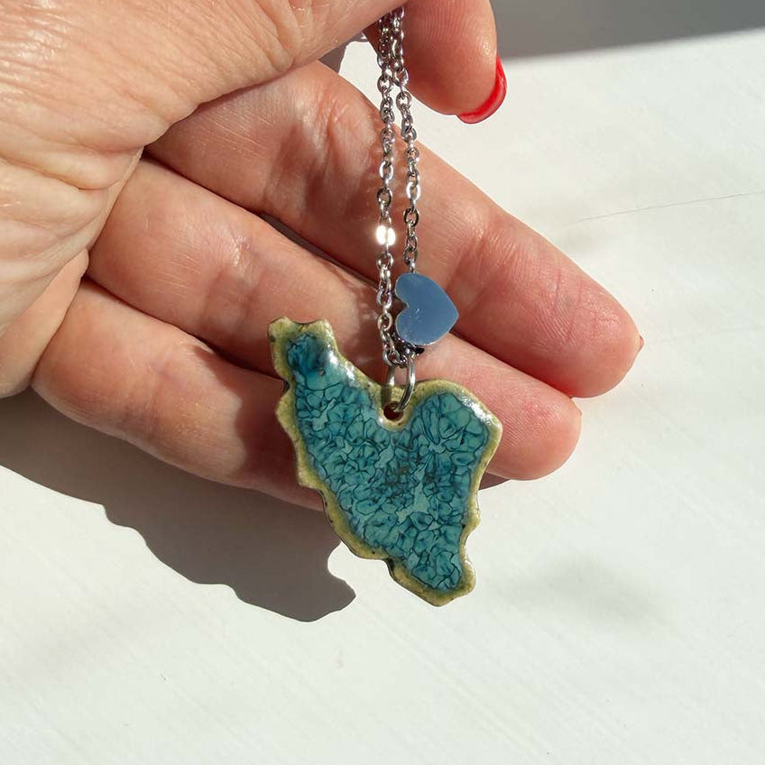 Handmade turquoise ceramic pendant shaped like the map of Iran, with a crackle reactive glaze and a small silver heart bead on a stainless steel chain, held in the palm of a hand.