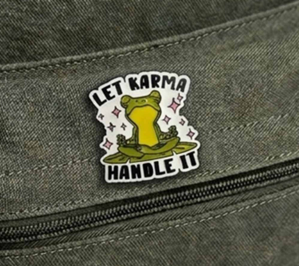 A close-up of a rectangular enamel pin attached to a dark olive green canvas zipper pouch. The pin features a cartoon frog meditating on a lily pad with the text 'LET KARMA HANDLE IT' in black lettering.