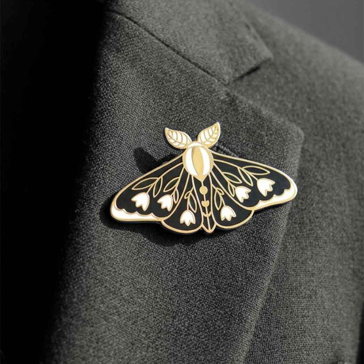 A gold and black moth-shaped lapel pin with white floral details, pinned to a dark charcoal gray suit jacket lapel.