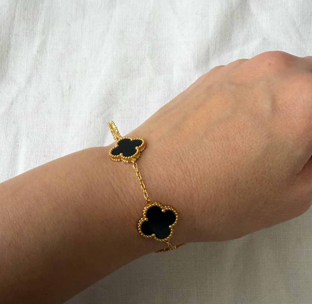 Gold bracelet with black clover-shaped charms on a wrist against a white background