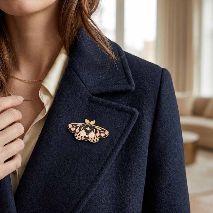 A close-up lifestyle shot of a woman wearing a navy blue wool coat over a cream shirt, featuring a detailed enamel moth brooch on the lapel.