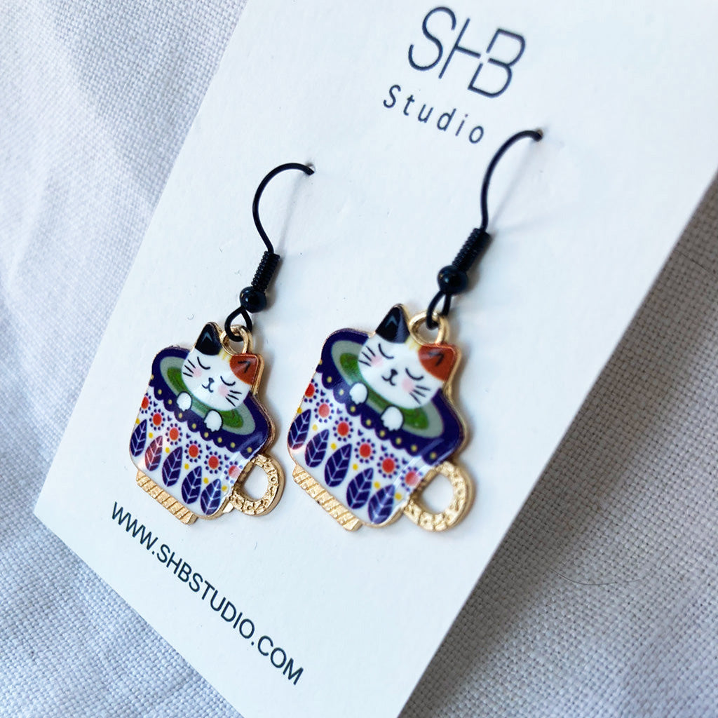 A pair of whimsical cat earrings in a teacup design with blue and white colors, displayed on a white background with &