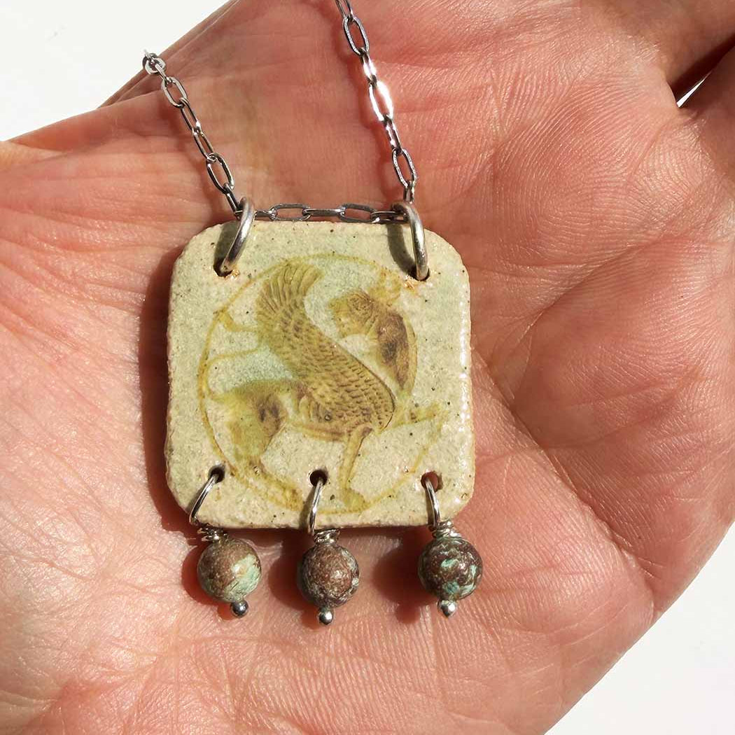 Close-up of a Persian Simorgh ceramic necklace in the palm of a hand, showing the sepia transfer of the mythological bird and turquoise bead drops.