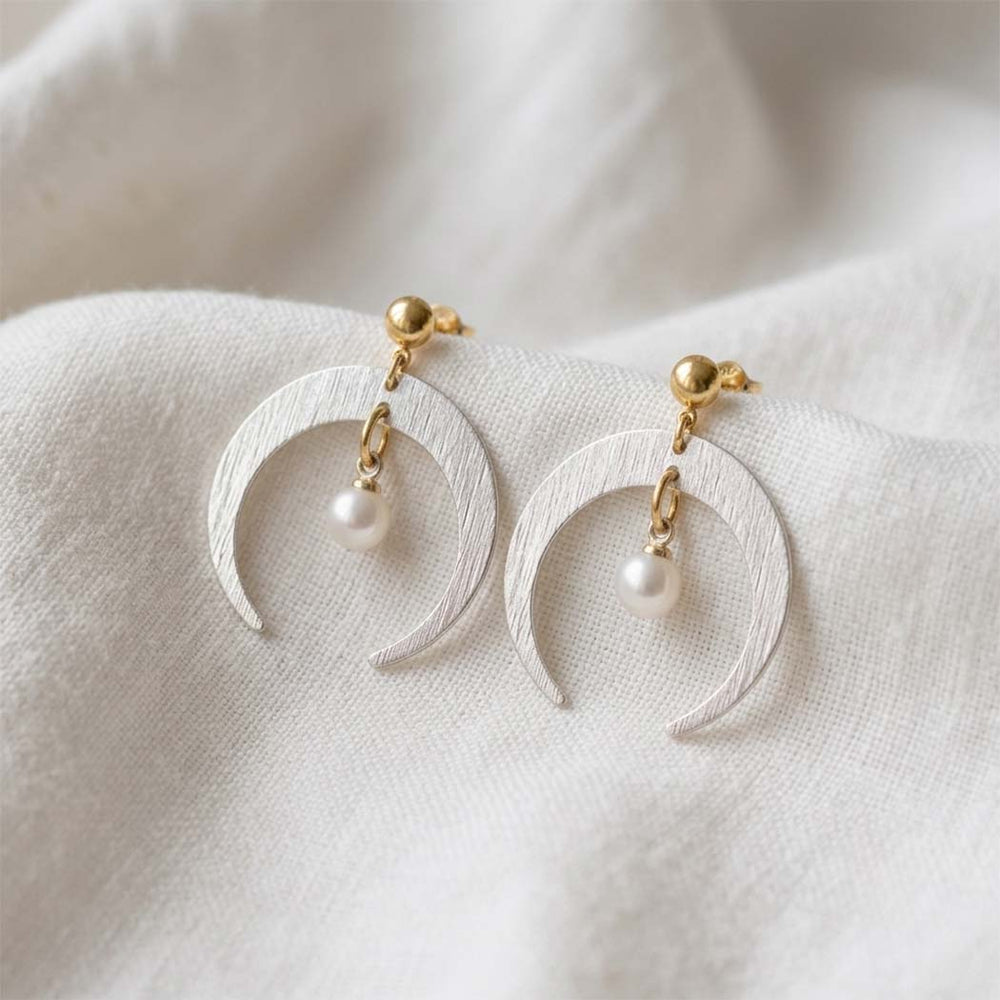 A pair of crescent moon drop earrings with textured white metal, gold posts, and small white pearl beads, resting on a soft white fabric background.