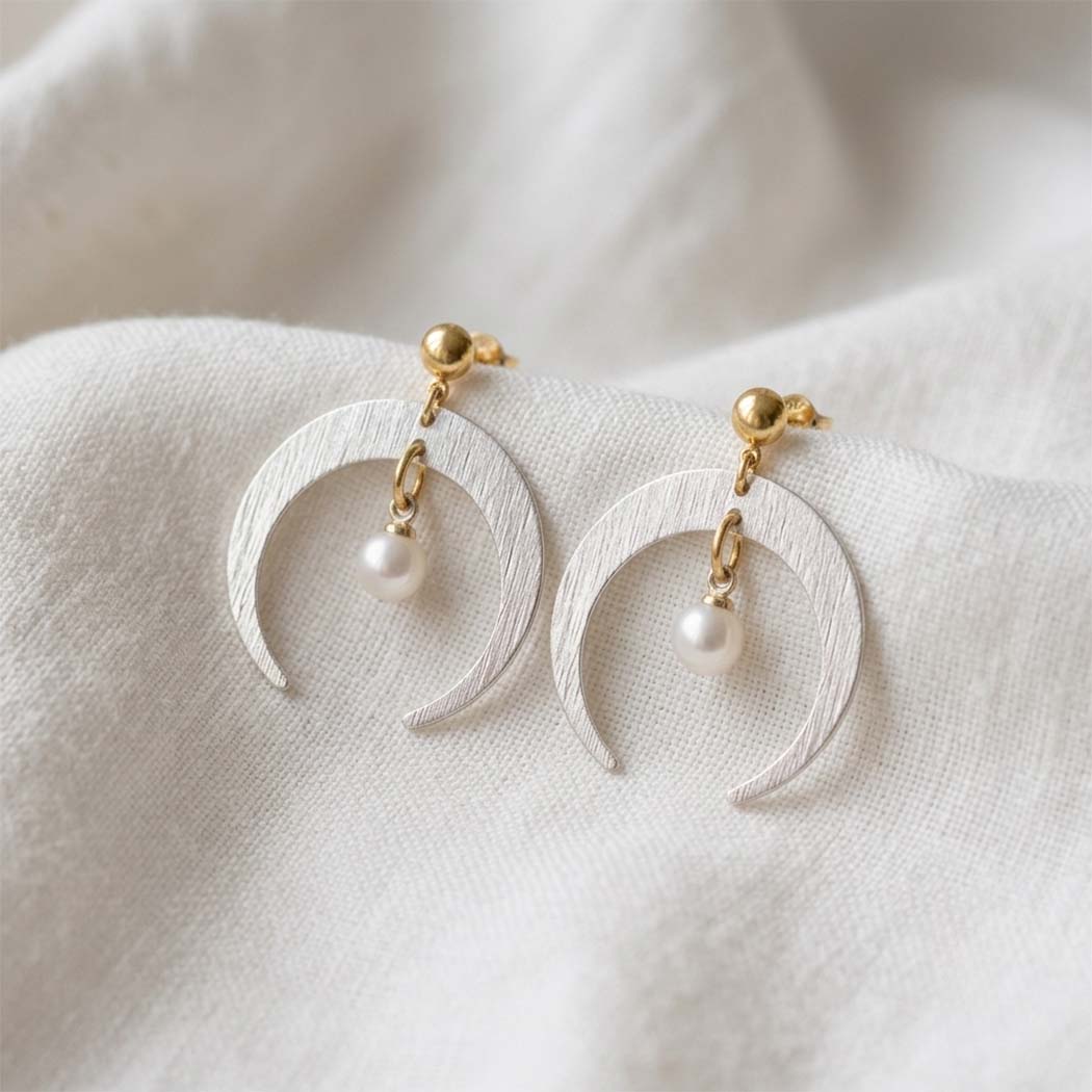 A pair of crescent moon drop earrings with textured white metal, gold posts, and small white pearl beads, resting on a soft white fabric background.