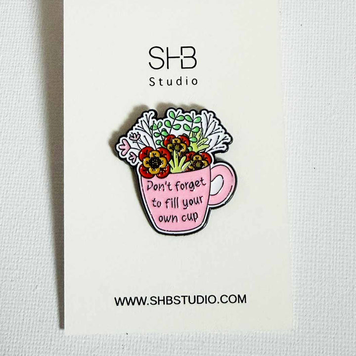 A pink enamel pin shaped like a mug filled with flowers and greenery, featuring the text &