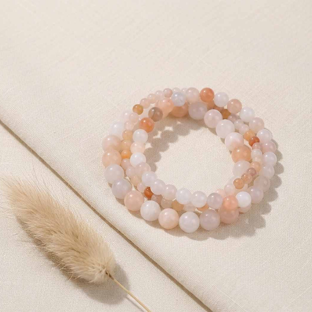 A set of three beaded stretch bracelets made of peach, pink, and white translucent round beads, arranged on a textured beige fabric background next to a dried bunny tail grass stem.