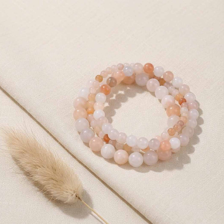 A set of three beaded stretch bracelets made of peach, pink, and white translucent round beads, arranged on a textured beige fabric background next to a dried bunny tail grass stem.