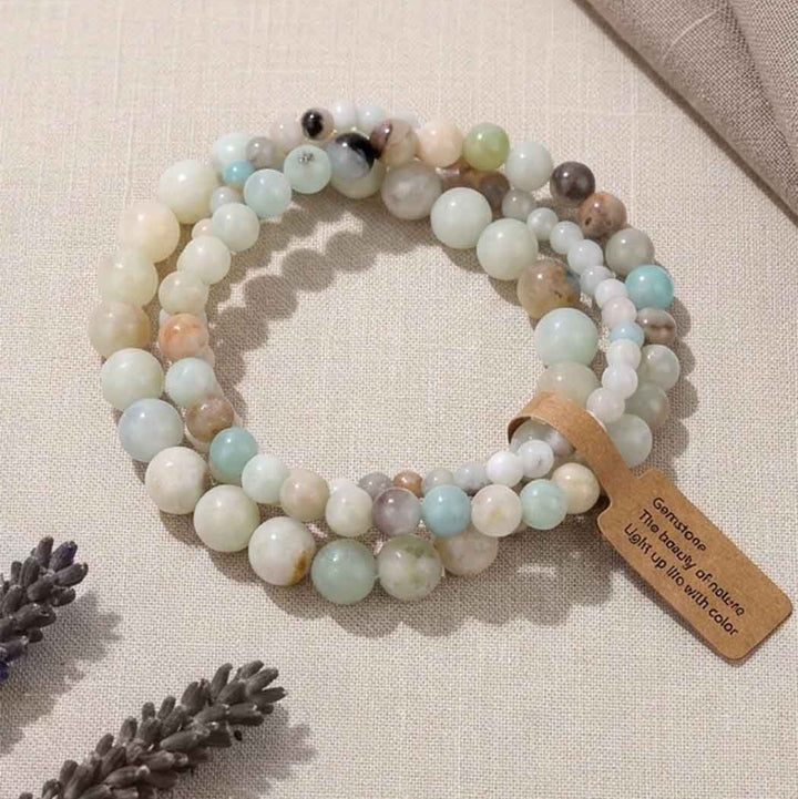 Three beaded stretch bracelets made of light-colored stone beads in pastel shades of mint, cream, peach, and gray, arranged on a beige fabric background with a small brown colour label attached.