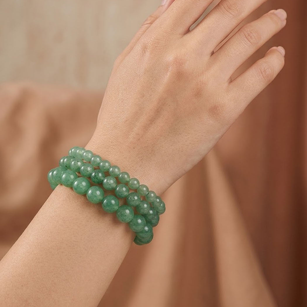 A close-up of a person's hand and wrist wearing three stacked green beaded bracelets against a soft, warm brown background.