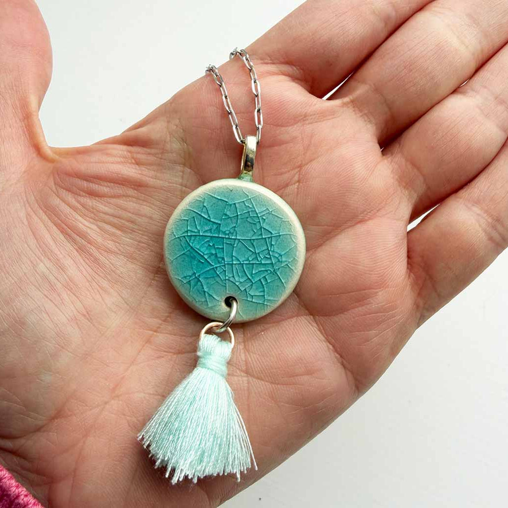 Turquoise ceramic pendant with tassel held in a hand against a white background