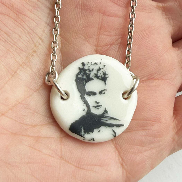 Necklace with a pendant featuring a black and white portrait Frida Kahlo Mexican painter non a white background, held in a hand.