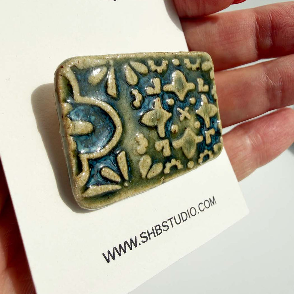 handmade brooch ceramic tile held by a hand on a card with SHB Studio branding.