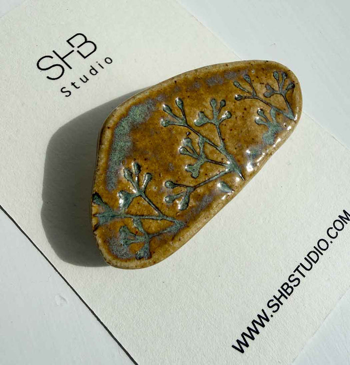 brooch with floral pattern on a white card with SHB Studio branding.