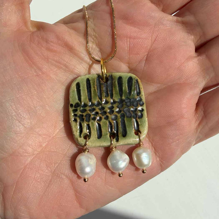 Hand holding a pendant with a green square design and pearl drops on a light background