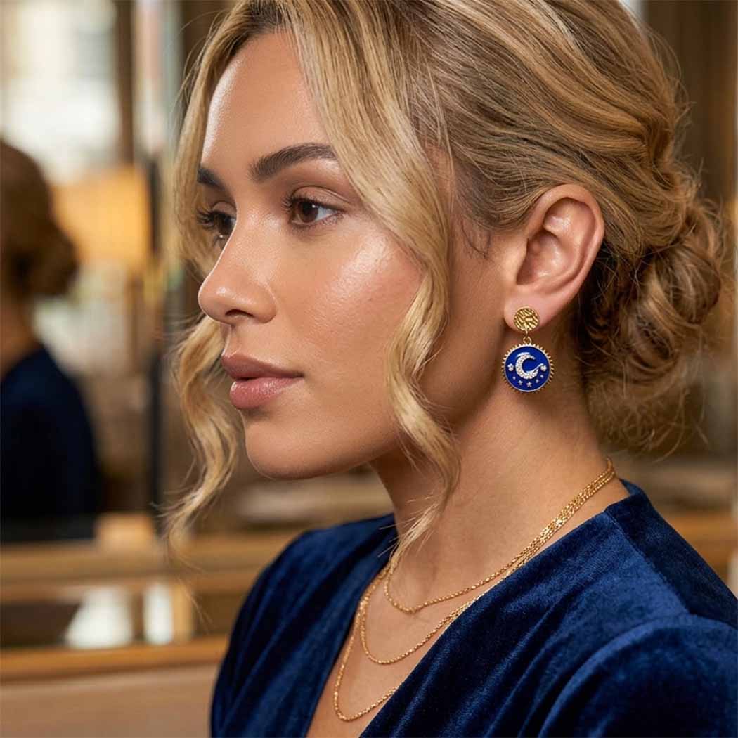A woman in profile wearing a gold-toned circular earring with a blue center and white crescent moon, paired with layered gold necklaces and a dark blue velvet top.