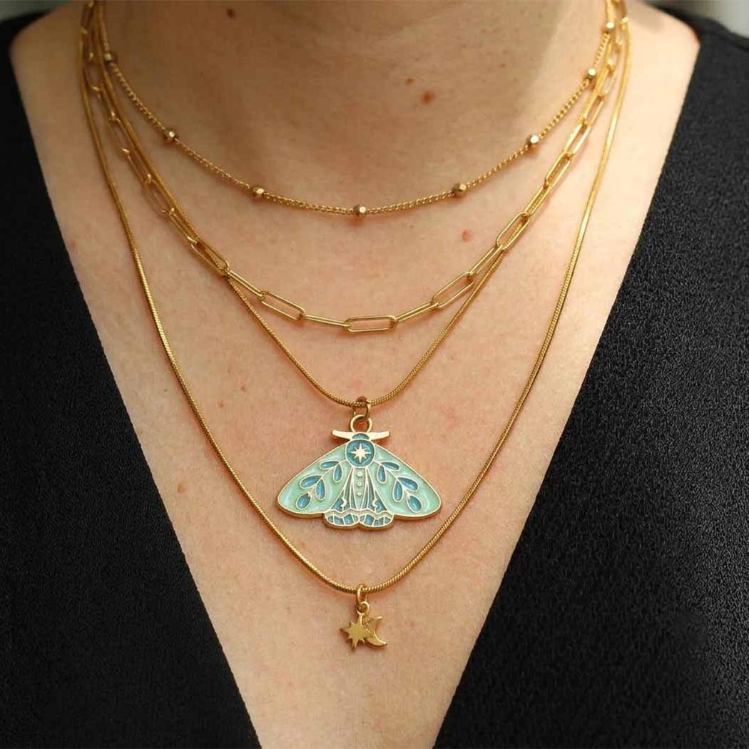 A close-up of a person wearing a layered gold necklace set with a light sage green and white moth pendant and a small gold star charm, paired with a black top.