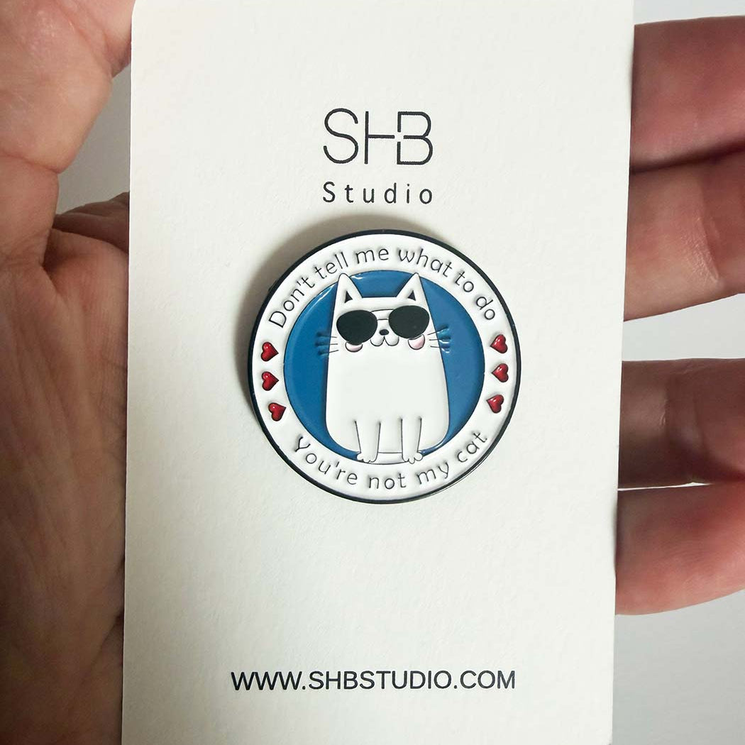 A circular enamel pin featuring a white cat with black sunglasses on a blue background, surrounded by the text &