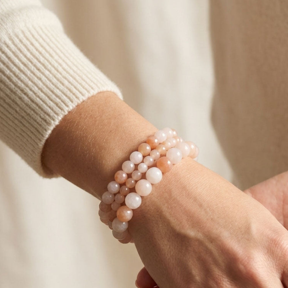 A close-up of a person's wrist wearing a double-strand beaded bracelet made of pale pink, peach, and white round beads, styled with a cream ribbed knit sweater.