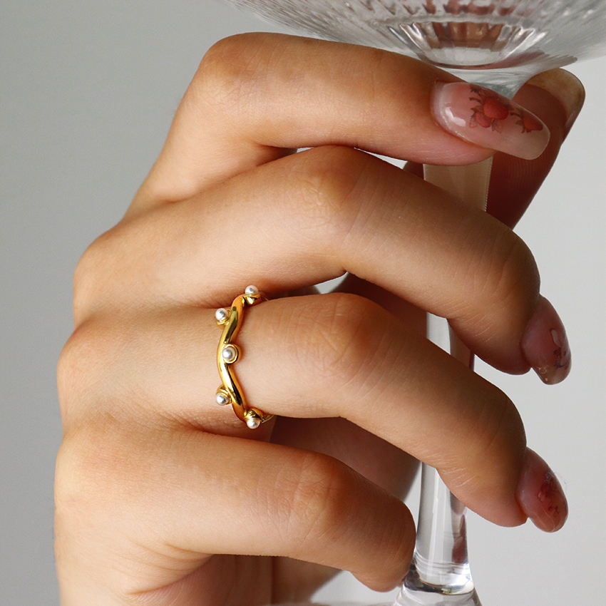 Hand wearing a gold ring with small pearls, holding a glass against a neutral background