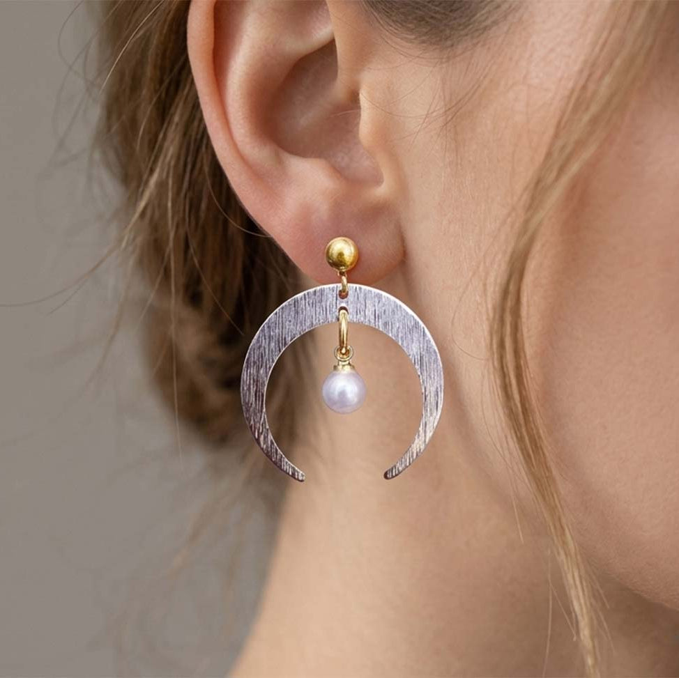 A close-up of a model's ear wearing a crescent moon dangle earring. The earring features a textured silver crescent moon with a small white pearl dangling from a gold-toned post.