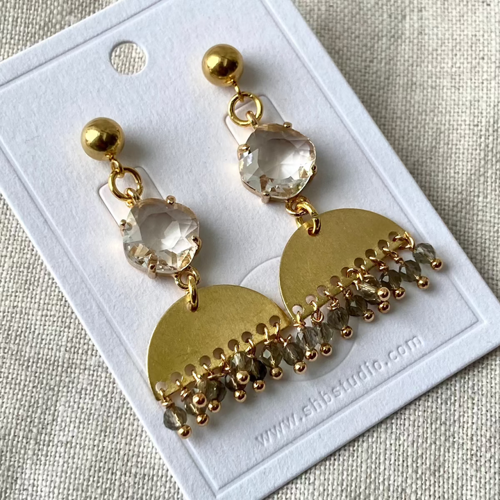 A pair of gold-plated brass earrings with large crystal balls and smaller crystal beads attached to them, displayed on a white card with the brand name &