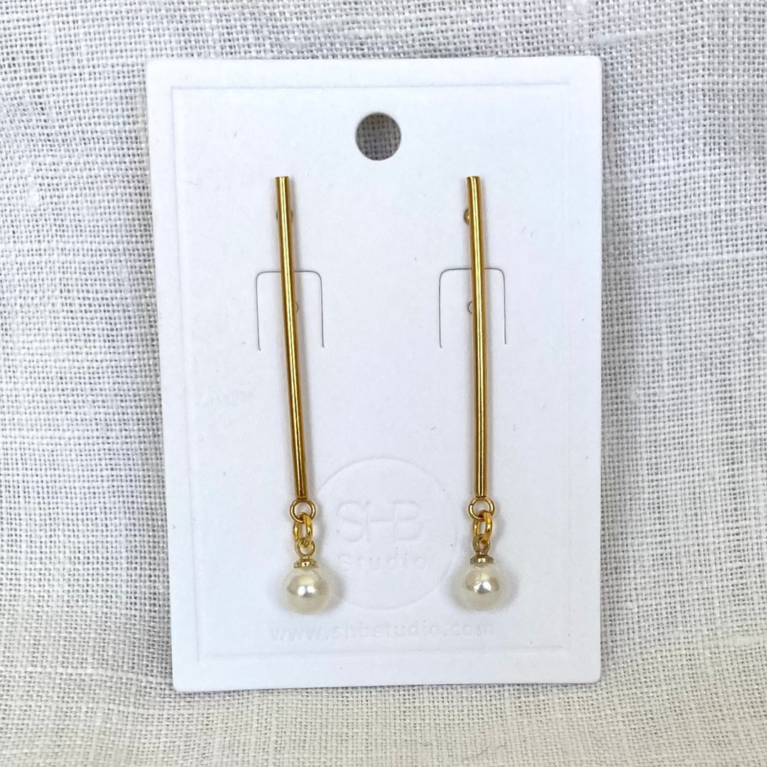 A pair of minimal long gold and pearl earrings displayed on a white card, with the pearl drops at the bottom of gold-plated rods.