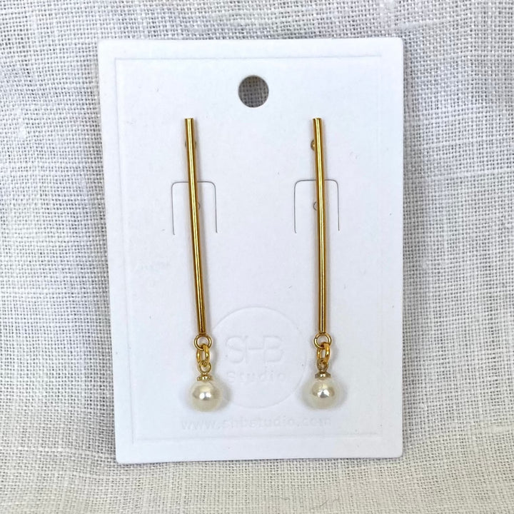 A pair of minimal long gold and pearl earrings displayed on a white card, with the pearl drops at the bottom of gold-plated rods.